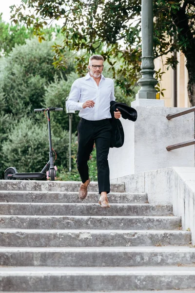 Businessman running down the stairs to catch his train at the station. Young financial manager running down the stairs talking with earphones and leaving the rental electric scooter on the sidewalk
