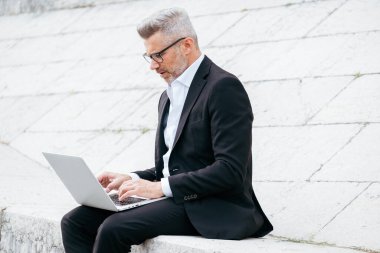 İş adamı şehir merkezinde merdivenlerde otururken dizüstü bilgisayarda çalışıyor.