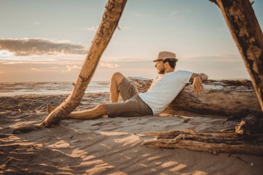 Yaz şapkasıyla gevşeyen genç bir adam çöl sahilindeki tahta kütüğün üzerinde gün batımına bakan bir birayla yatıyordu.