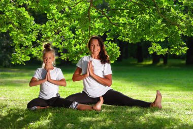 Anne ve küçük kızı birlikte yoga ya da pilates spor alıştırması yapıyorlar yaz parkında çimenler üzerinde ve gülümsüyorlar. Aile sporu ve aktivitesi kavramı. Sağlıklı yaşam tarzı.