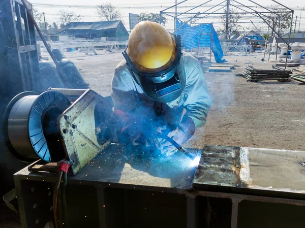 Kaynakçı, Flux Cored Arc Welding (FCAW) işlemiyle bir çelik yapı çalışması yapıyor ve endüstriyel fabrikada güvenlik için kişisel koruyucu ekipman (PPE) ile uygun şekilde giyiniyor.