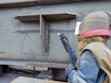 İşçiler, çeliğin yüzeyini boyamadan önce yüzey profili oluşturmak için kaynak yüzeyi ve çelik yapıyı patlatıyor..