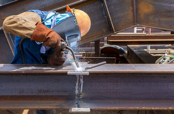 Kaynakçı, çelik yapı çalışması için Flux Cored Arc Resding (FCAW) işlemine ekleme yapıyor ve endüstriyel fabrikada güvenlik için kişisel koruyucu ekipman (PPE) ile uygun giyiniyor.