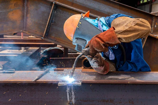 Kaynakçı, çelik yapı çalışması için Flux Cored Arc Resding (FCAW) işlemine ekleme yapıyor ve endüstriyel fabrikada güvenlik için kişisel koruyucu ekipman (PPE) ile uygun giyiniyor.
