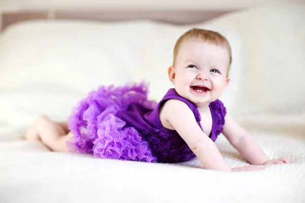 Smějící se holčička v purpurových šatech na bílé posteli — Stock fotografie