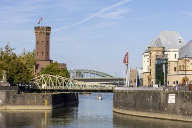 Köln ve Ren Nehri 'nin manzarası Çikolata Müzesi' nin yanında sergilenir ve tekneler parıldayan suyun üzerinde yavaşça süzülür. Modern binalar ve köprüler, parlak ve berrak bir gökyüzü altında tarihi bir cazibeyle harmanlanır..
