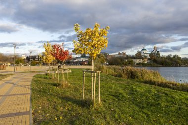Güneşli sonbahar havasında nehir boyunca uzanan bir yaya yolu. Altın yapraklar toprağı kaplıyor, ağaçlar ılık ışıkta parıldıyor, ve hafif esinti huzurlu havayı huzur ve güzellikle dolduruyor..