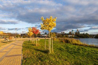 Güneşli sonbahar havasında nehir boyunca uzanan bir yaya yolu. Altın yapraklar toprağı kaplıyor, ağaçlar ılık ışıkta parıldıyor, ve hafif esinti huzurlu havayı huzur ve güzellikle dolduruyor..