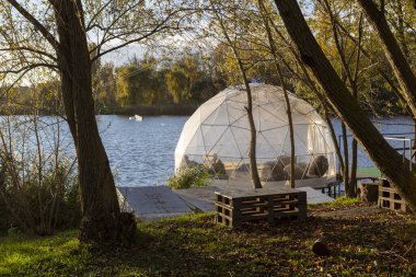 Güneşli bir sonbahar gününde, altın ağaçlar ve sakin suyla çevrili şeffaf bir kubbe nehir kıyısında duruyor. İçeride, yumuşak ışık ve sıcaklık dinlenmek, düşünmek ve dinlenmek için huzurlu bir alan yaratır..