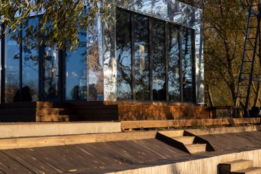 Sonbahar renkleriyle çevrili bir parkın ortasında cam ve ayna bir bina duruyor. Altın yapraklar parlak yüzeyini yansıtır. Mimariyle doğayı sakin, uyumlu ve zarif bir mevsimlik manzarayla harmanlar..