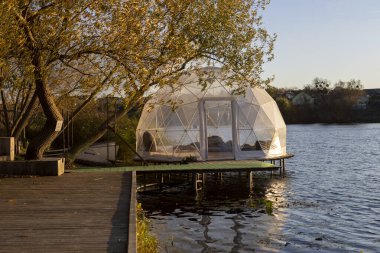 Güneşli bir sonbahar gününde, altın ağaçlar ve sakin suyla çevrili şeffaf bir kubbe nehir kıyısında duruyor. İçeride, yumuşak ışık ve sıcaklık dinlenmek, düşünmek ve dinlenmek için huzurlu bir alan yaratır..