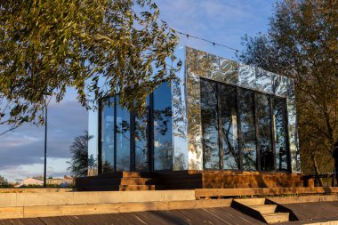 Sonbahar renkleriyle çevrili bir parkın ortasında cam ve ayna bir bina duruyor. Altın yapraklar parlak yüzeyini yansıtır. Mimariyle doğayı sakin, uyumlu ve zarif bir mevsimlik manzarayla harmanlar..