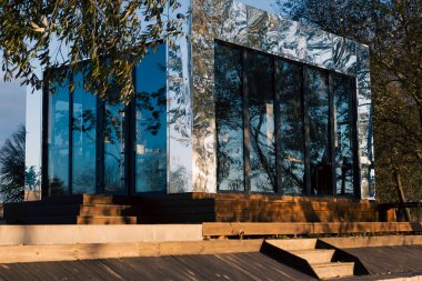 Sonbahar renkleriyle çevrili bir parkın ortasında cam ve ayna bir bina duruyor. Altın yapraklar parlak yüzeyini yansıtır. Mimariyle doğayı sakin, uyumlu ve zarif bir mevsimlik manzarayla harmanlar..