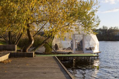 Güneşli bir sonbahar gününde, altın ağaçlar ve sakin suyla çevrili şeffaf bir kubbe nehir kıyısında duruyor. İçeride, yumuşak ışık ve sıcaklık dinlenmek, düşünmek ve dinlenmek için huzurlu bir alan yaratır..