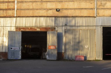 Geniş açık kapıları olan endüstriyel bir garaj. Beton zemin, metal yapılar ve tozlu pencerelerden süzülen yumuşak ışıklarla donatılmış geniş bir iç mekan..