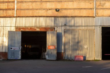 Geniş açık kapıları olan endüstriyel bir garaj. Beton zemin, metal yapılar ve tozlu pencerelerden süzülen yumuşak ışıklarla donatılmış geniş bir iç mekan..