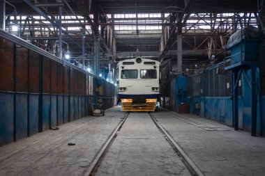 Elektrik treni, atölyedeki makineler, aletler ve endüstriyel ekipmanlarla çevrili bir inşaat ve onarım tesisinin önüne park edilmiştir..