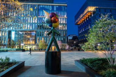 Modern cam ve çelik mimarisi olan bir iş merkezinin akşam manzarası pencerelere yansıyan yumuşak ışıklar ve yakınındaki renkli çiçek tarhlı bir meydan..