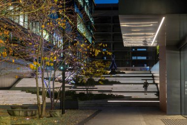 Modern cam ve çelik mimarisi olan bir iş merkezinin akşam manzarası pencerelere yansıyan yumuşak ışıklar ve yakınındaki renkli çiçek tarhlı bir meydan..