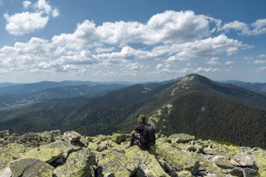 Sırt çantası ve şapkası olan bir turist, bulutlu bir yaz gününde bir dağ kayasının üzerinde oturur ve uzaklara bakar..
