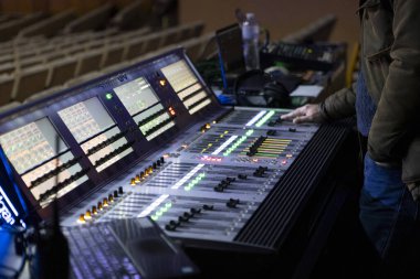 Bir ses mühendisi konser sırasında dijital karıştırma konsolunda çalışır, canlı sahne ışıkları kontrol yüzeyini aydınlatırken seviyeleri ve efektleri ayarlar ve canlı ses üretiminin dinamik bir sahnesini yaratır..