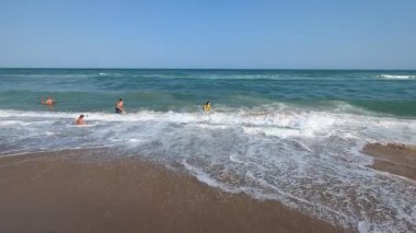 Yumuşak dalgalar kumlu sahile yuvarlanıyor, sıcak öğleden sonra güneşi altında parlıyor ve huzurlu, parlak bir sahil atmosferi yaratıyor..