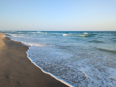 Deniz yumuşak, ritmik dalgalar oluşturur. Sakin bir öğleden sonra ışığı ve berrak bir gökyüzü tarafından aydınlatılır..