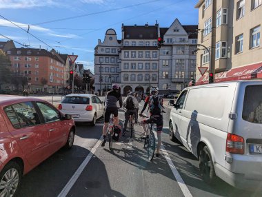 Köln, Almanya, 1 Ekim 2025: Bisikletçiler yoğun bir Köln yolunda kırmızı ışıkta bekliyorlar. Şehir binaları, sokak tabelaları ve gün ışığı sürücüler rotalarına devam etmeden önce duraklarken canlı bir şehir atmosferi yaratıyor..