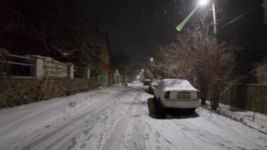 Akşamları, evler ve park edilmiş arabalarla dolu, karla kaplı sessiz bir cadde. Pencerelerden ve sokak lambalarından gelen yumuşak ışık taze karı yansıtıyor ve sakin bir kış manzarası yaratıyor..