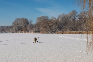 Yalnız bir balıkçı, açık bir kış gününde karla kaplı donmuş bir nehirde sabırla oturur. Parlak güneş ışığı ve uçsuz bucaksız beyaz manzara yalnızlık ve geleneksel kış balıkçılığının sakin bir atmosferini yaratır..