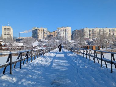 Donmuş bir gölet ve açık mavi bir gökyüzünün altında ağır şekilde karla kaplı ahşap bir köprü içeren nefes kesici bir kış manzarası. Parlak güneş ışığı karların parlamasını sağlıyor, doğada dingin ve el değmemiş bir atmosfer yaratıyor..