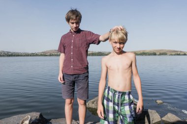 Çocuklar gölün kıyısında duruyordu. Gölün yanında iki arkadaş. Çocuk yüzme şortuyla gölün kenarında duruyor. Çocuk güneşleniyor..