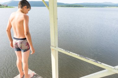 Çocuk suya atlamak için kulede dinleniyor. Çocuk göl kenarında dinleniyor. Şortlu bir yüzücü. Yaz tatili. Genç güneş banyosu. Büyük bir yükseklikten suya atlamak. Yüksek irtifa korkusu