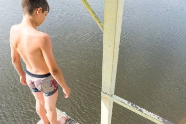 Çocuk suya atlamak için kulede dinleniyor. Çocuk göl kenarında dinleniyor. Şortlu bir yüzücü. Yaz tatili. Genç güneş banyosu. Büyük bir yükseklikten suya atlamak. Yüksek irtifa korkusu