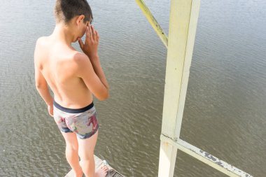 Çocuk suya atlamak için kulede dinleniyor. Çocuk göl kenarında dinleniyor. Şortlu bir yüzücü. Yaz tatili. Genç güneş banyosu. Büyük bir yükseklikten suya atlamak. Yüksek irtifa korkusu