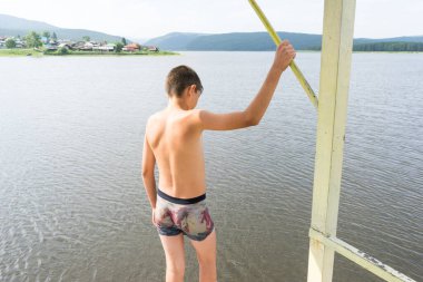 Çocuk suya atlamak için kulede dinleniyor. Çocuk göl kenarında dinleniyor. Şortlu bir yüzücü. Yaz tatili. Genç güneş banyosu. Büyük bir yükseklikten suya atlamak. Yüksek irtifa korkusu