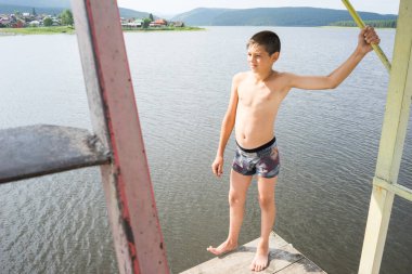 Çocuk suya atlamak için kulede dinleniyor. Çocuk göl kenarında dinleniyor. Şortlu bir yüzücü. Yaz tatili. Genç güneş banyosu. Büyük bir yükseklikten suya atlamak. Yüksek irtifa korkusu