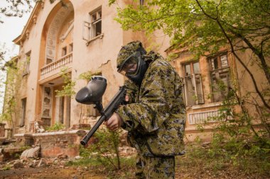 Paintball oynayan genç adam. Kamuflaj kıyafetli çocuk paintball silahını doğrultuyor. Koruyucu giysili, maskeli çocuk..