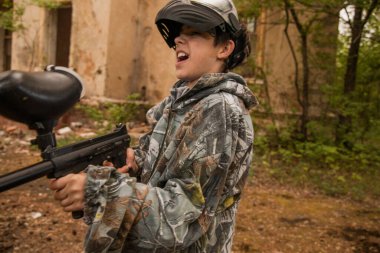 Paintball oynayan genç adam. Kamuflaj kıyafetli çocuk paintball silahını doğrultuyor. Koruyucu giysili, maskeli çocuk..