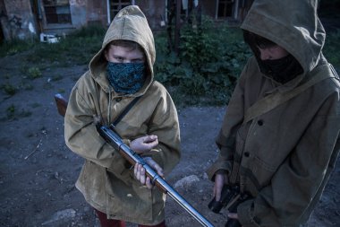 Koruyucu kapüşonlu ceketli iki çocuk terk edilmiş bir binanın önünde duruyor. Silahlı bir çocuk..