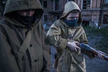 Koruyucu kapüşonlu ceketli iki çocuk terk edilmiş bir binanın önünde duruyor. Silahlı bir çocuk..