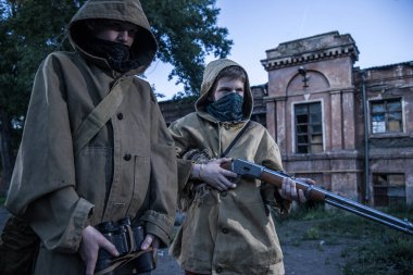 Koruyucu kapüşonlu ceketli iki çocuk terk edilmiş bir binanın önünde duruyor. Silahlı bir çocuk..