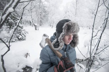 Şapkalı ve kalın elbiseli bir çocuk elinde bir balta tutuyor. Genç bir adam kış ormanında..