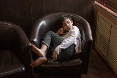 Beyaz gömlekli ve kot pantolonlu bir çocuk sandalyede oturuyor. Bir kafede dinlenen bir genç.