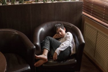 Beyaz gömlekli ve kot pantolonlu bir çocuk sandalyede oturuyor. Bir kafede dinlenen bir genç.