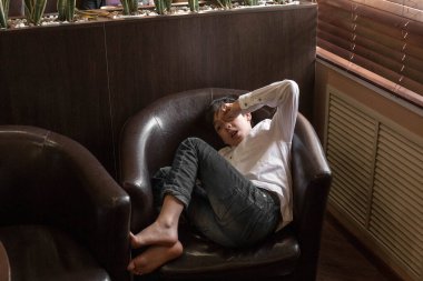 Beyaz gömlekli ve kot pantolonlu bir çocuk sandalyede oturuyor. Bir kafede dinlenen bir genç.