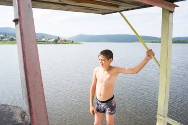 Çocuk suya atlamak için kulede dinleniyor. Çocuk göl kenarında dinleniyor. Şortlu bir yüzücü. Yaz tatili. Genç güneş banyosu. Büyük bir yükseklikten suya atlamak. Yüksek irtifa korkusu