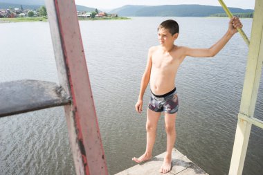 Çocuk suya atlamak için kulede dinleniyor. Çocuk göl kenarında dinleniyor. Şortlu bir yüzücü. Yaz tatili. Genç güneş banyosu. Büyük bir yükseklikten suya atlamak. Yüksek irtifa korkusu