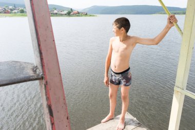 Çocuk suya atlamak için kulede dinleniyor. Çocuk göl kenarında dinleniyor. Şortlu bir yüzücü. Yaz tatili. Genç güneş banyosu. Büyük bir yükseklikten suya atlamak. Yüksek irtifa korkusu