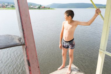 Çocuk suya atlamak için kulede dinleniyor. Çocuk göl kenarında dinleniyor. Şortlu bir yüzücü. Yaz tatili. Genç güneş banyosu. Büyük bir yükseklikten suya atlamak. Yüksek irtifa korkusu